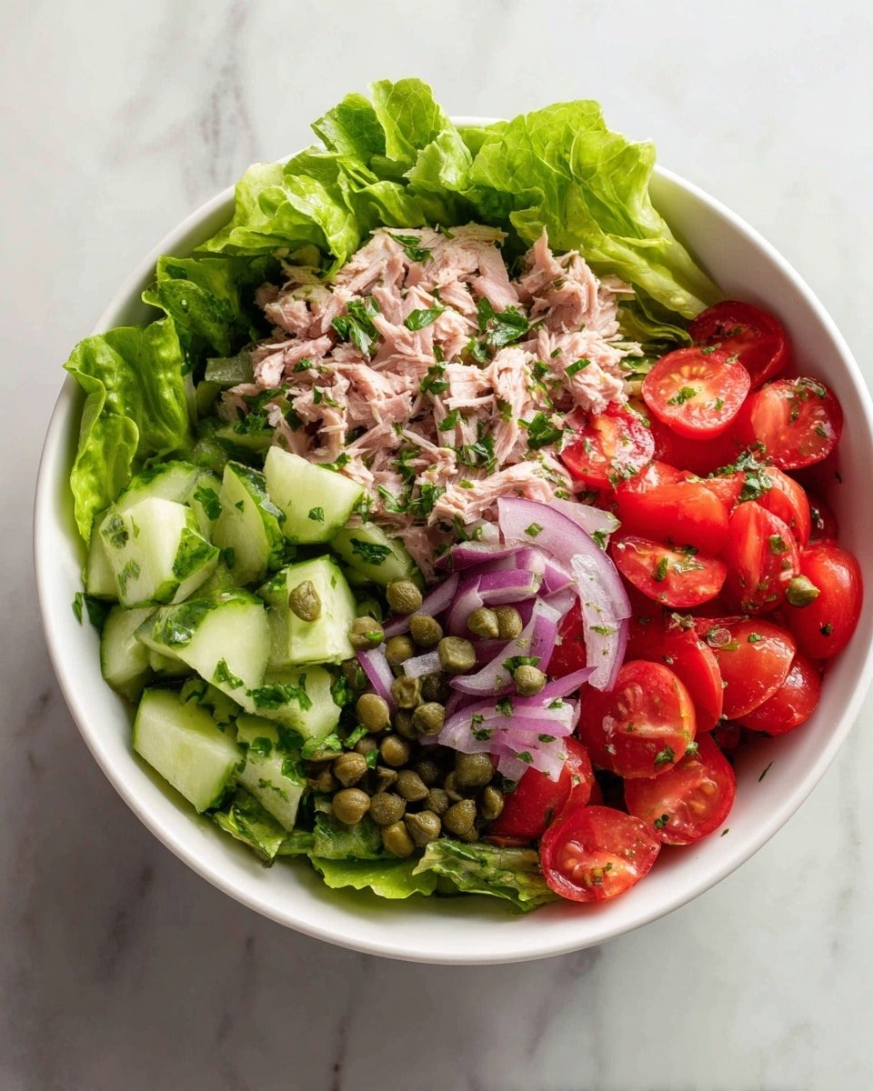 A white bowl filled with a three-layer salad placed on a white marbled surface. The bottom layer is made of green leafy lettuce with a fresh, crisp texture. The middle layer contains halved bright red cherry tomatoes and light green cucumber chunks. The top layer is a mix of light pink shredded tuna, chopped red onions, small green capers, and sprinkled green herbs. The bowl is slightly close-up, showing the fresh and colorful ingredients clearly. Photo taken with an iphone --ar 4:5 --v 7 — Mediterranean Tuna Salad with Avocado & Capers, healthy tuna salad, easy Mediterranean salad, lemon tuna salad, protein-packed tuna salad