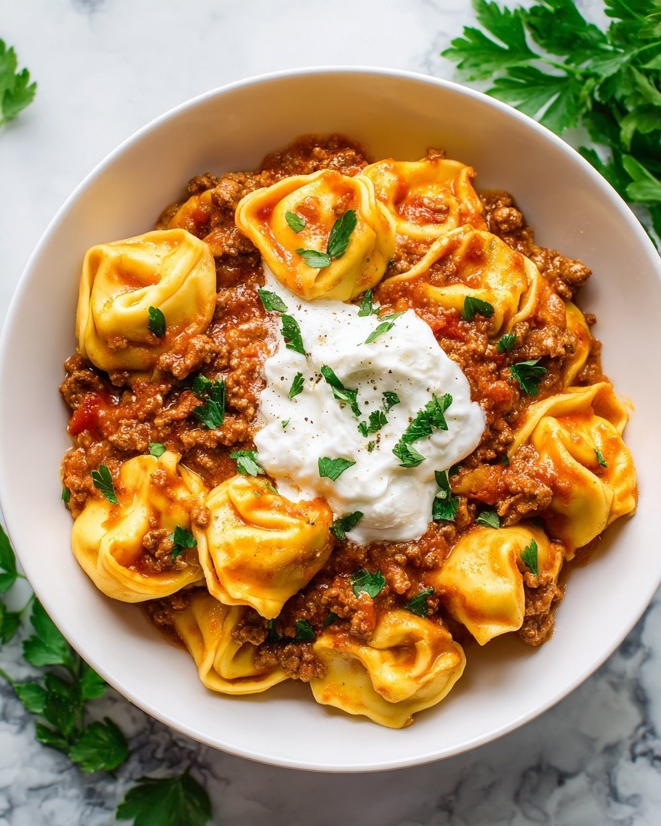 In the white bowl, there is a rich, thick tomato sauce mixed with ground meat, covering around two layers of tortellini pasta that are pale yellow and softly curled. On top of the sauce and pasta, in the middle, is a generous dollop of smooth white cream, giving a creamy contrast. Scattered over the whole dish are bright green cilantro leaves adding fresh color. The bowl rests on a white marbled surface. photo taken with an iphone --ar 4:5 --v 7 — Cheesy Beef Enchilada Tortellini, enchilada tortellini recipe, cheesy beef pasta dish, comfort food pasta, easy weeknight dinner