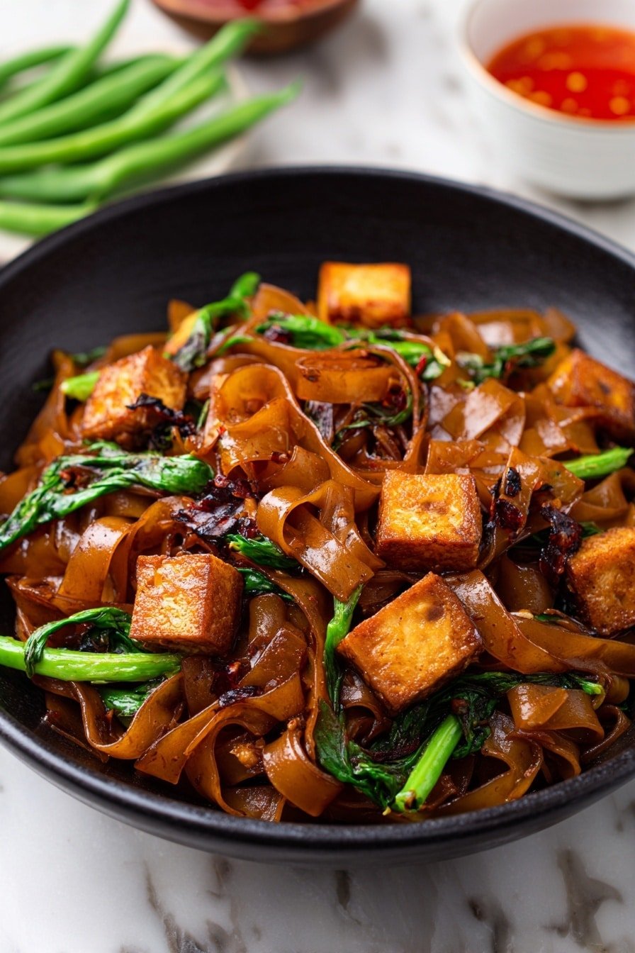 A black bowl filled with wide, glossy, brown stir-fried noodles layered with small, golden-brown tofu cubes and bright green vegetable pieces scattered throughout. The noodles appear soft and slightly shiny, mixed with dark leafy bits and small chili flakes, giving a tasty, spicy look. The bowl sits on a white marbled surface, with some green beans and a white bowl with red sauce blurred in the background. photo taken with an iphone --ar 2:3 --v 7 — Thai Drunken Noodles, Thai Drunken Noodles recipe, easy Thai noodle stir-fry, flavorful Thai noodle dish, quick Thai dinner ideas