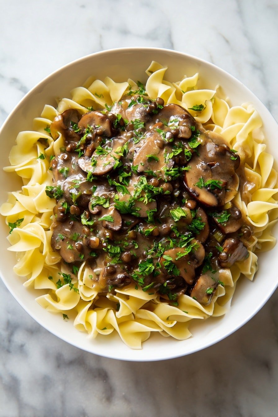 A white bowl filled with wide, flat egg noodles forming the bottom layer, their pale yellow color and slightly ruffled edges visible around the sides. On top, a thick layer of creamy mushroom sauce covers the noodles, with visible large slices of cooked brown mushrooms and smaller lentils mixed into the sauce. The sauce is rich brown and shiny, contrasting with the noodles. Bright green chopped parsley is sprinkled evenly over the top, adding a fresh pop of color. The bowl sits on a white marbled surface. Photo taken with an iphone --ar 2:3 --v 7 — Lentil Mushroom Stroganoff, vegan mushroom stroganoff, hearty vegetarian stroganoff, creamy lentil mushroom sauce, comforting mushroom and lentil dish