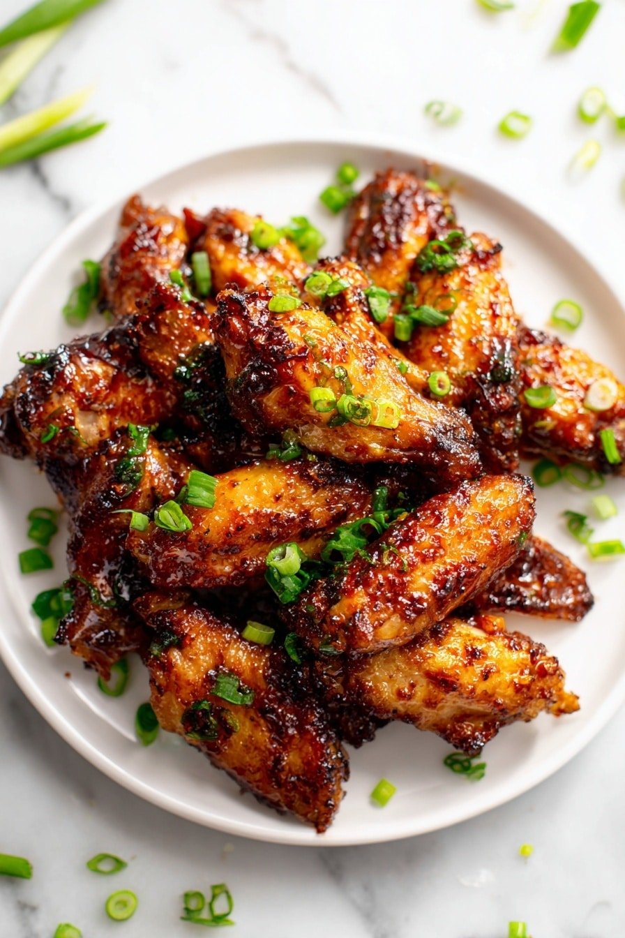 A round white plate filled with a single layer of shiny, golden-brown chicken wings coated in a sticky sauce with a slightly charred look. Small bright green chopped scallions are scattered on top, adding fresh color and contrast. The plate sits on a white marbled surface with some green scallion pieces near it. The skin of the wings looks crispy with visible spices on the glossy sauce. photo taken with an iphone --ar 2:3 --v 7 — Hot Honey Chicken, spicy honey chicken, crispy honey glazed chicken, baked hot honey chicken, easy spicy chicken recipe