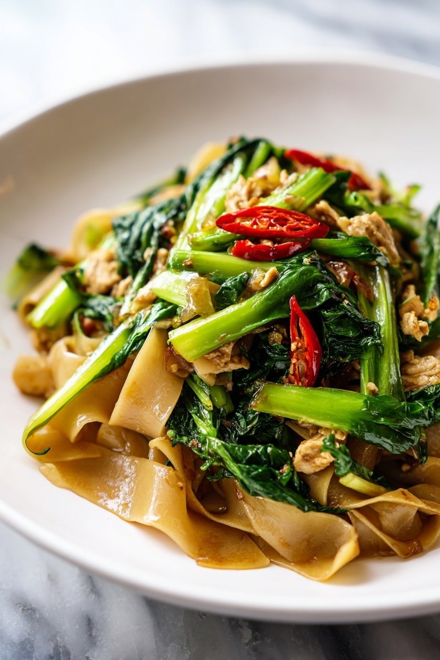 A white plate filled with wide, flat brown noodles coated in a shiny dark sauce forms the base layer. Mixed throughout are bright green leafy vegetables with thick stems, adding a fresh and vibrant look. Scattered on top are small pieces of browned meat, offering a textured contrast. Thin slices of red chili peppers are spread evenly across the dish, adding a pop of bright color. The dish sits on a white marbled surface, capturing the glossy and rich appearance of the noodles and greens. photo taken with an iphone --ar 2:3 --v 7 — Easy Drunken Noodles, Thai drunken noodles recipe, flavorful stir-fry with basil, quick Asian noodle dish, homemade drunken noodles