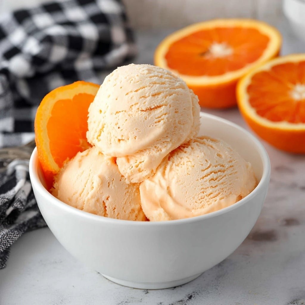 The image shows three scoops of light orange ice cream stacked in a white bowl with a smooth texture, slightly creamy and soft-looking with small uneven edges. The bowl is placed on a gray surface with a black spoon nearby, and in the background, there is a blurred half orange fruit and a white bowl with a wooden utensil slightly visible. The overall setting is bright with a white marbled texture at the back. photo taken with an iphone --ar 1:1 --v 7 — Homemade Orange Ice Cream, orange ice cream recipe, citrus ice cream, homemade frozen dessert, easy orange ice cream