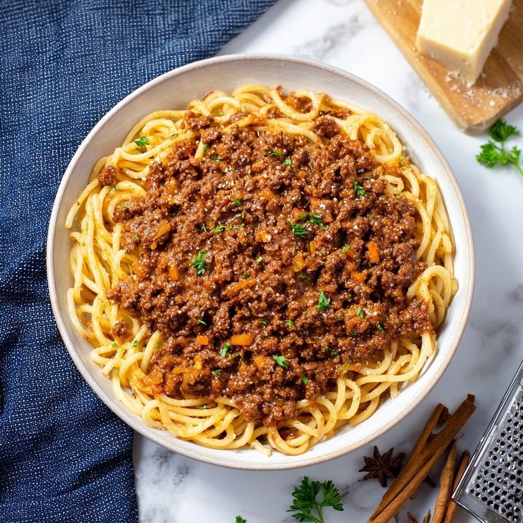 A white shallow bowl is filled with thick, round spaghetti noodles underneath a generous layer of chunky brown meat sauce mixed with small pieces of orange and green vegetables. The sauce covers most of the noodles but some are visible twisting underneath. Small green herb pieces are sprinkled lightly on top, adding a touch of color. The bowl sits on a white marbled surface with a blue textured cloth to the left and a block of grated cheese and metal grater slightly out of focus on the right. Cinnamon sticks and whole cloves are scattered loosely near the bottom. Photo taken with an iphone --ar 1:1 --v 7 — Greek Meat Sauce with Bucatini, Greek-inspired meat sauce, hearty Greek pasta, Mediterranean meat sauce, flavorful Greek pasta dish