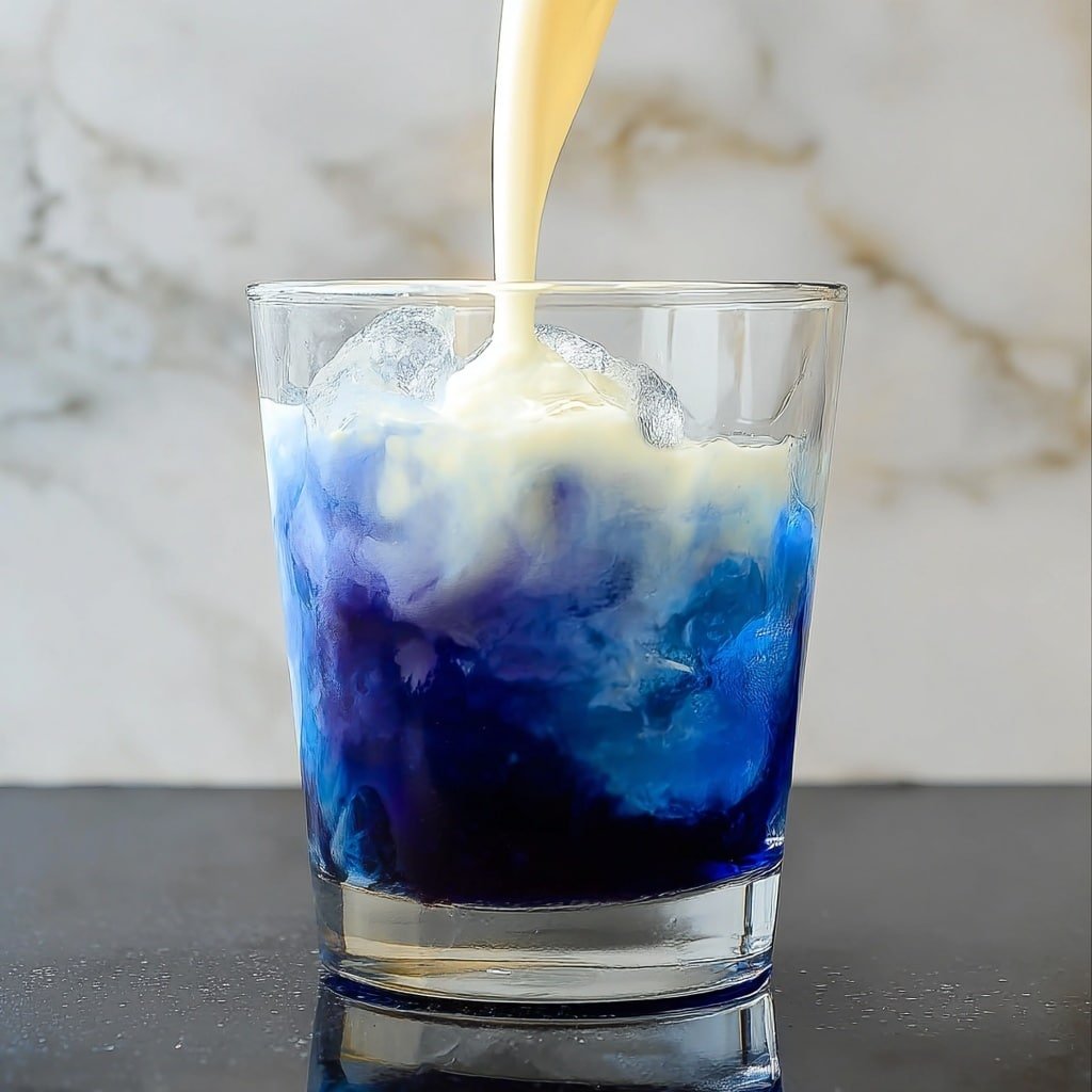 A clear glass filled with a dark blue liquid at the bottom layer with ice cubes inside. On top of the blue layer, a creamy white liquid is being poured, creating a swirling effect where the two colors mix softly. The glass sits on a dark surface that reflects the drink slightly, with a white marbled background behind. photo taken with an iphone --ar 1:1 --v 7 — Blue Butterfly Pea Latte, Blue Butterfly Pea Flower Drink, Healthy Butterfly Pea Latte, Vibrant Blue Latte Recipe, Floral Butterfly Pea Tea Latte