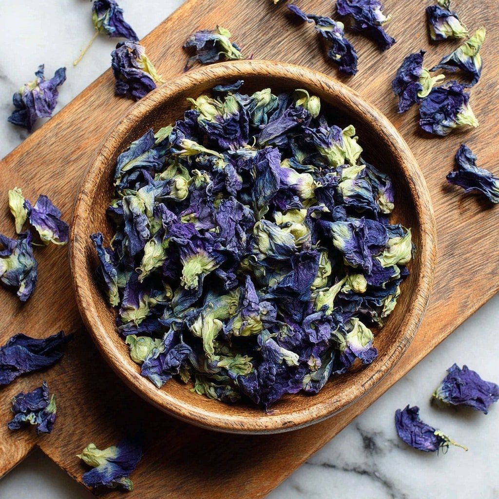 The image shows a round wooden bowl filled with dried butterfly pea flowers, which have deep purple petals with light green parts near the base. Some dried flowers are scattered around the bowl on a white marbled surface and a wooden board underneath. The petals have a rough, crinkled texture, and the colors contrast between the dark purple and pale green. The bowl is centered in the image, and the scattered flowers add a natural, casual feel. photo taken with an iphone --ar 1:1 --v 7 — Blue Butterfly Pea Latte, Blue Butterfly Pea Flower Drink, Healthy Butterfly Pea Latte, Vibrant Blue Latte Recipe, Floral Butterfly Pea Tea Latte