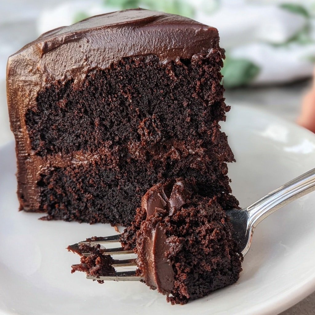 A thick slice of dark chocolate cake with two visible layers, rich and moist in texture, sits on a white plate. Each chocolate cake layer has a dense crumb, and between them is a thick layer of smooth, shiny dark chocolate frosting. The outside of the cake is fully covered with the same glossy dark chocolate frosting that drips slightly onto the plate. In the background, the remaining whole cake is visible on another white plate, with the same rich dark chocolate frosting covering it smoothly. The setup is on a white marbled surface with vibrant pink flowers and greenery blurred in the background. Photo taken with an iphone --ar 1:1 --v 7 — Brick Street Chocolate Cake, easy chocolate cake recipe, moist chocolate cake, rich chocolate cake, classic chocolate cake