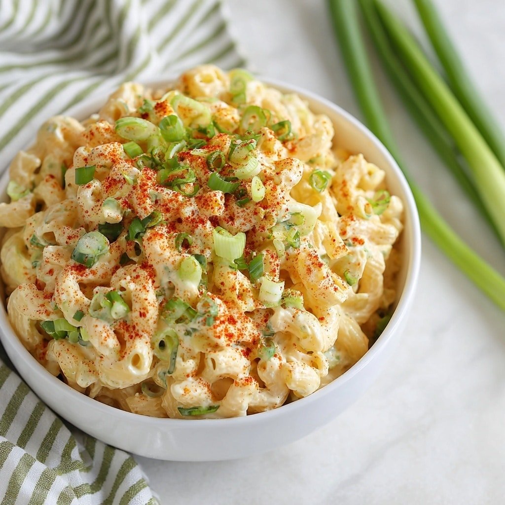 A white bowl filled with creamy macaroni salad made from elbow pasta covered in a pale yellow sauce. The top layer is sprinkled with bright green chopped scallions and a dusting of red paprika, adding color contrast. The bowl sits on a white marbled surface with green onions and a blurred striped cloth in the background. The macaroni pieces look soft and coated evenly in the sauce, making the dish look fresh and tasty. Photo taken with an iphone --ar 1:1 --v 7 — Creamy Deviled Egg Pasta Salad, deviled egg pasta salad, easy pasta salad recipes, quick picnic salads, flavorful pasta salad ideas