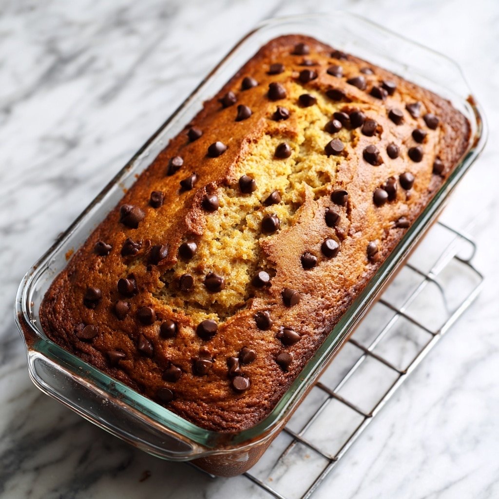 A golden brown baked loaf with a slightly rough and cracked texture, filled evenly with small dark brown chocolate chips scattered across the top and within. It sits inside a clear glass rectangular baking dish, placed on the middle rack of an oven with metal grates. The loaf looks soft and thick, about one solid layer rising just above the edges of the dish. The background is a white marbled texture. photo taken with an iphone --ar 1:1 --v 7 — High-Protein Banana Bread, healthy banana bread with protein powder, easy banana bread recipe, moist banana bread, nutritious breakfast bread